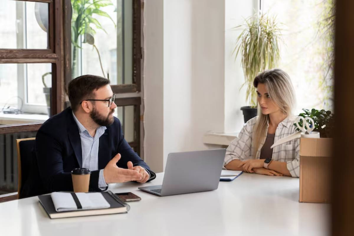 A business meeting in a bright, modern office with a laptop on the table, coffee cup, and plants visible in the background.