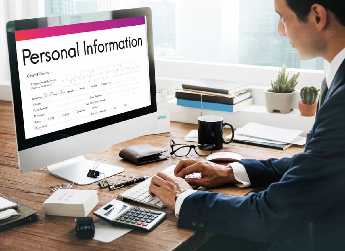 A person in a suit types on a keyboard, with a computer displaying a personal information form and office supplies nearby.