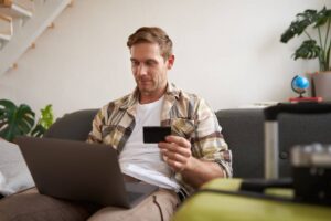 A person sits on a couch with a laptop open, holding a credit card, surrounded by plants and travel luggage.