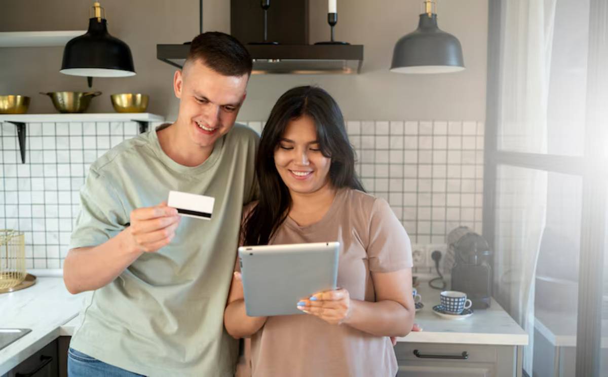 A couple stands in a modern kitchen, one holding a credit card and the other a tablet, both focused on their tasks. 