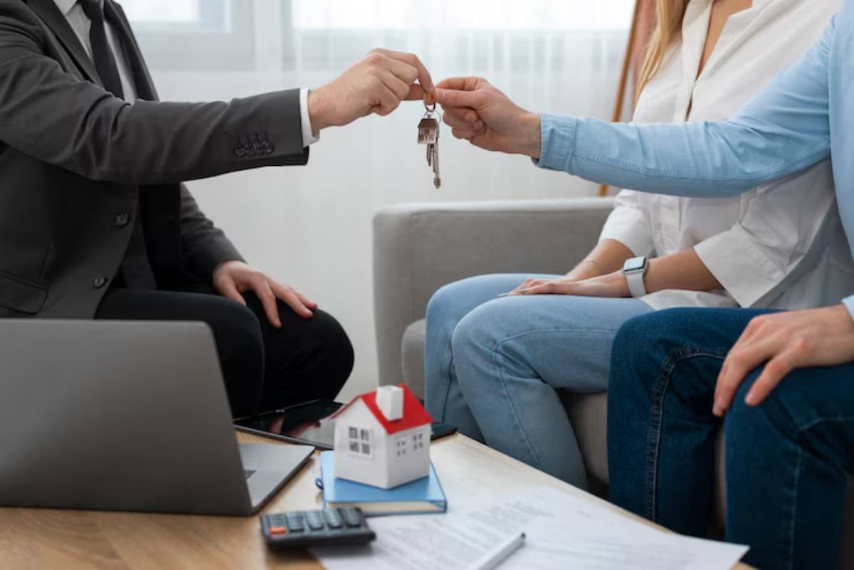 A real estate agent hands over house keys to a couple sitting on a couch, with documents and a miniature house on the table.