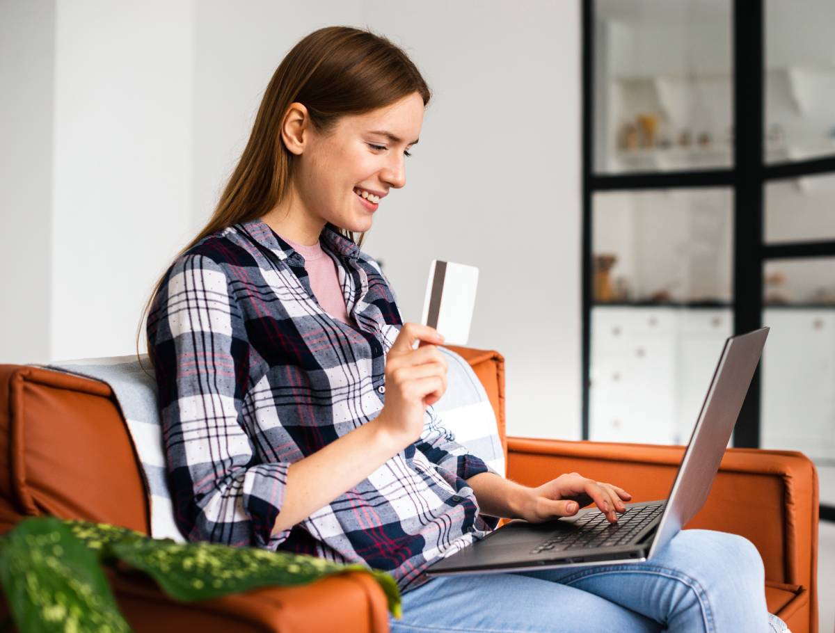 A person sits on a couch, working on a laptop while holding a credit card, surrounded by indoor plants and modern decor. 