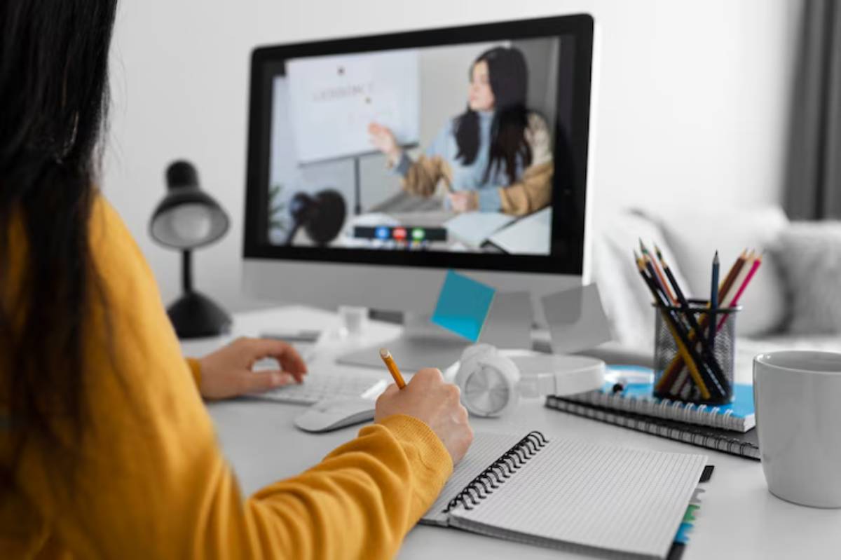 A person in a yellow sweater takes notes while participating in an online meeting on a desktop computer surrounded by stationery and a mug. 