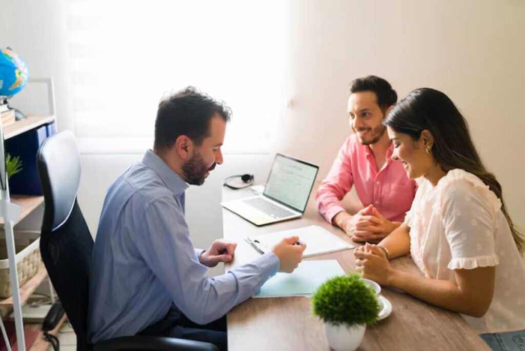 A man in a blue shirt sits at a desk, discussing with a couple, with a laptop and a small plant on the table in a bright office setting.