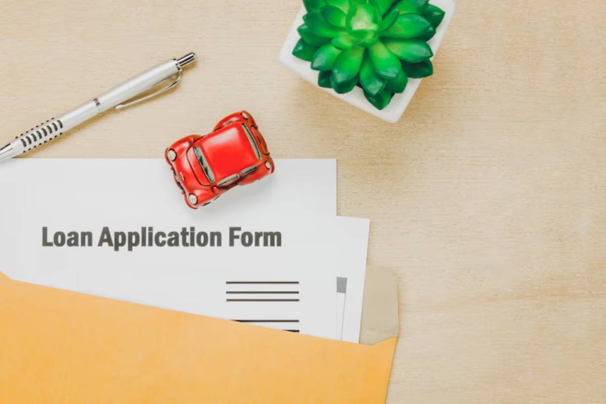 A red toy car rests on a loan application form, beside a silver pen and a small green succulent plant on a wooden table.