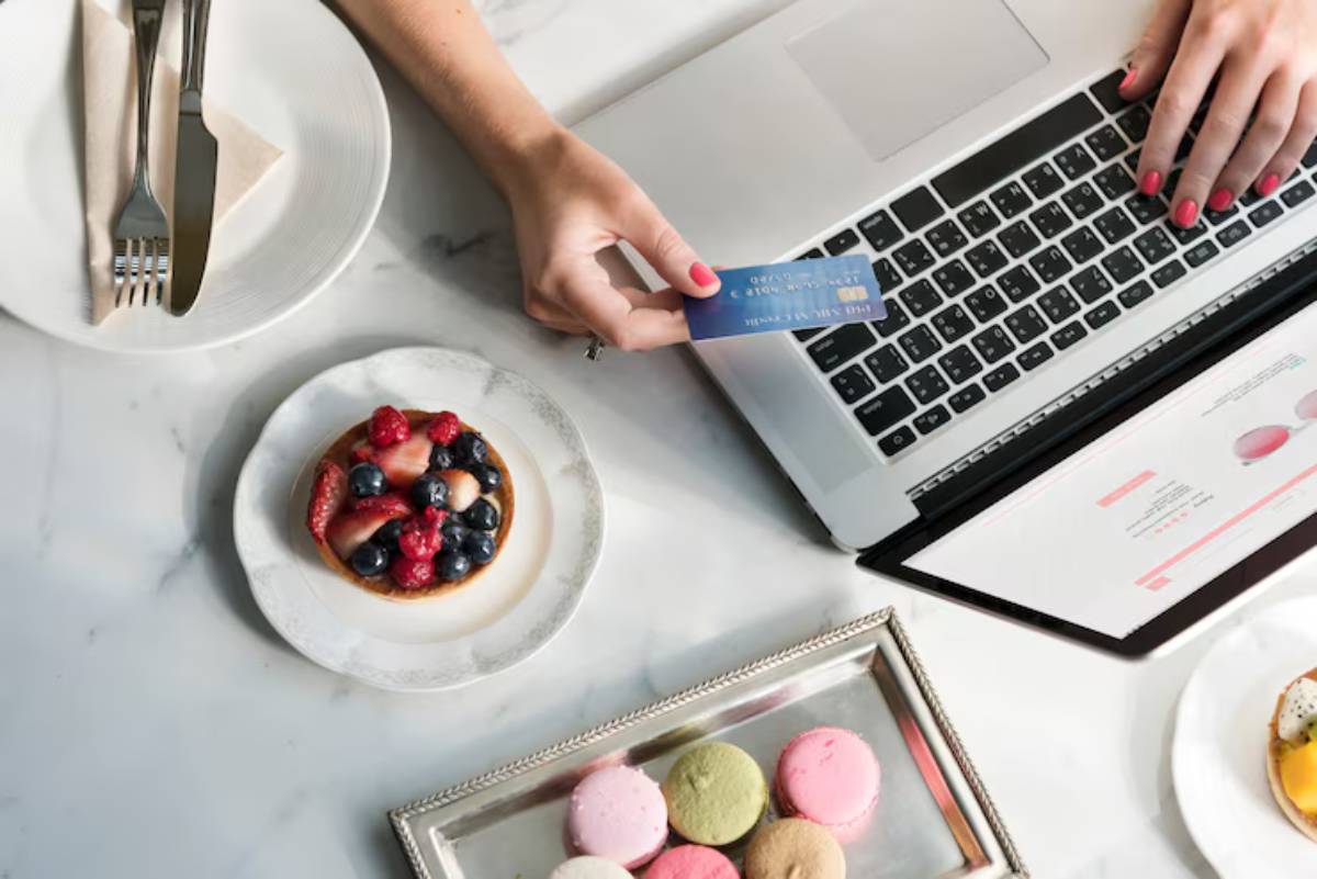 A hand holds a credit card above a laptop, next to a plate of fruit tart and colorful macarons on a marble table.