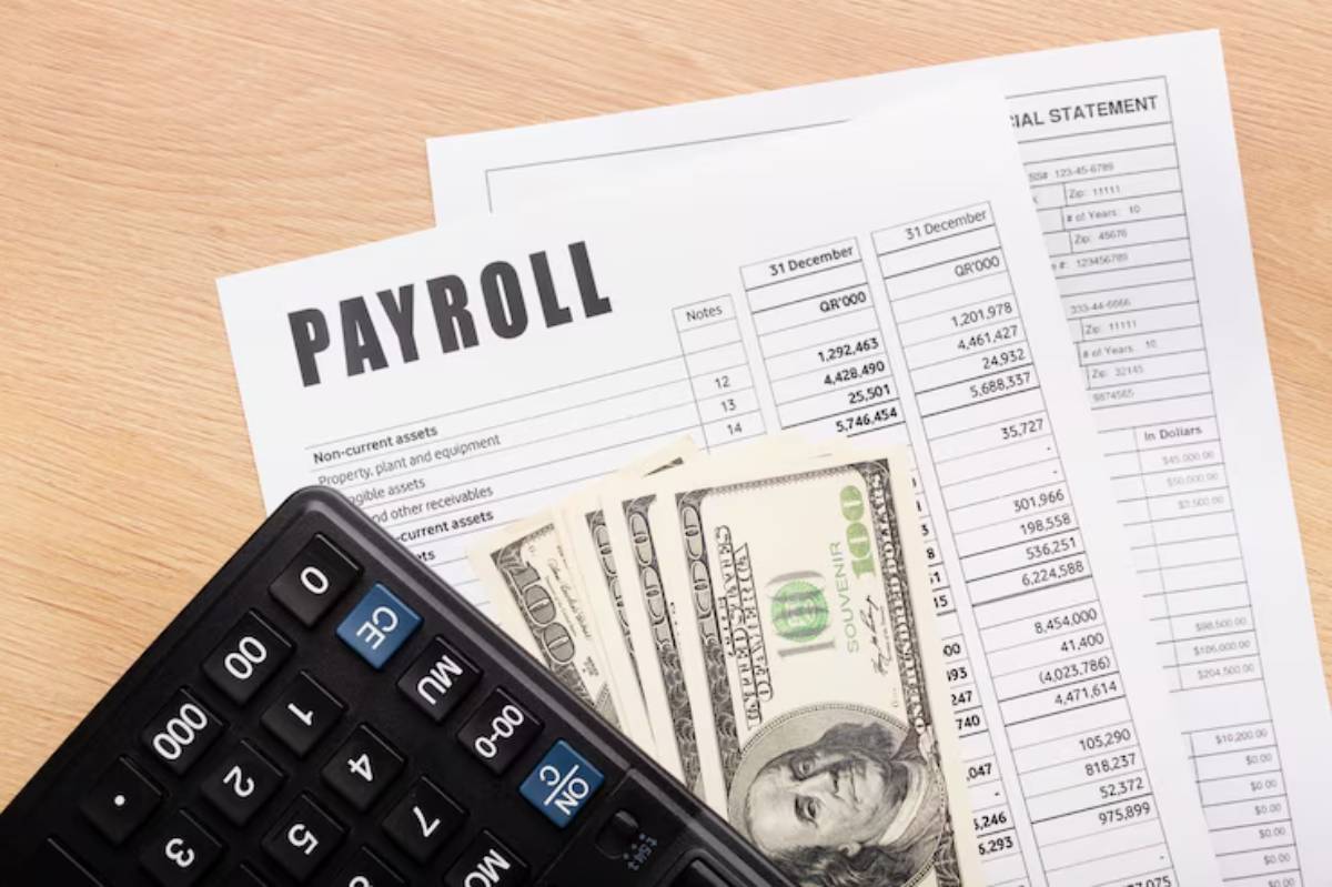 A calculator, cash, and payroll documents are arranged on a wooden surface, highlighting financial management tasks.