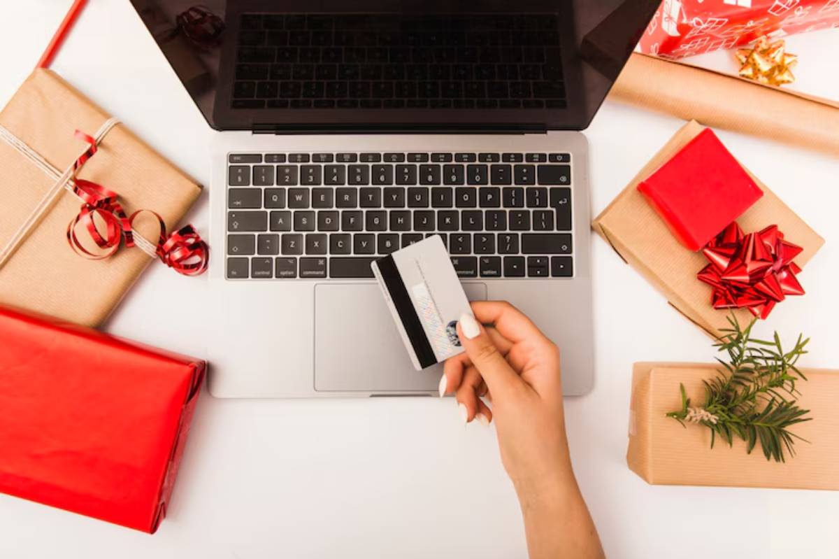 A hand holds a credit card above a laptop keyboard, surrounded by festive wrapped gifts and decorations.  