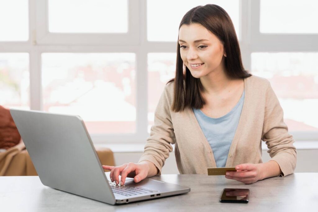 A person sits at a table using a laptop, holding a credit card in one hand, with a smartphone nearby and bright windows in the background.