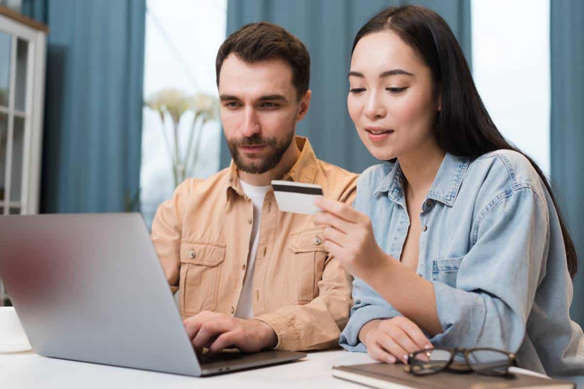 A man and woman are seated at a table, using a laptop, with the woman holding a credit card, focused on their online activity.
