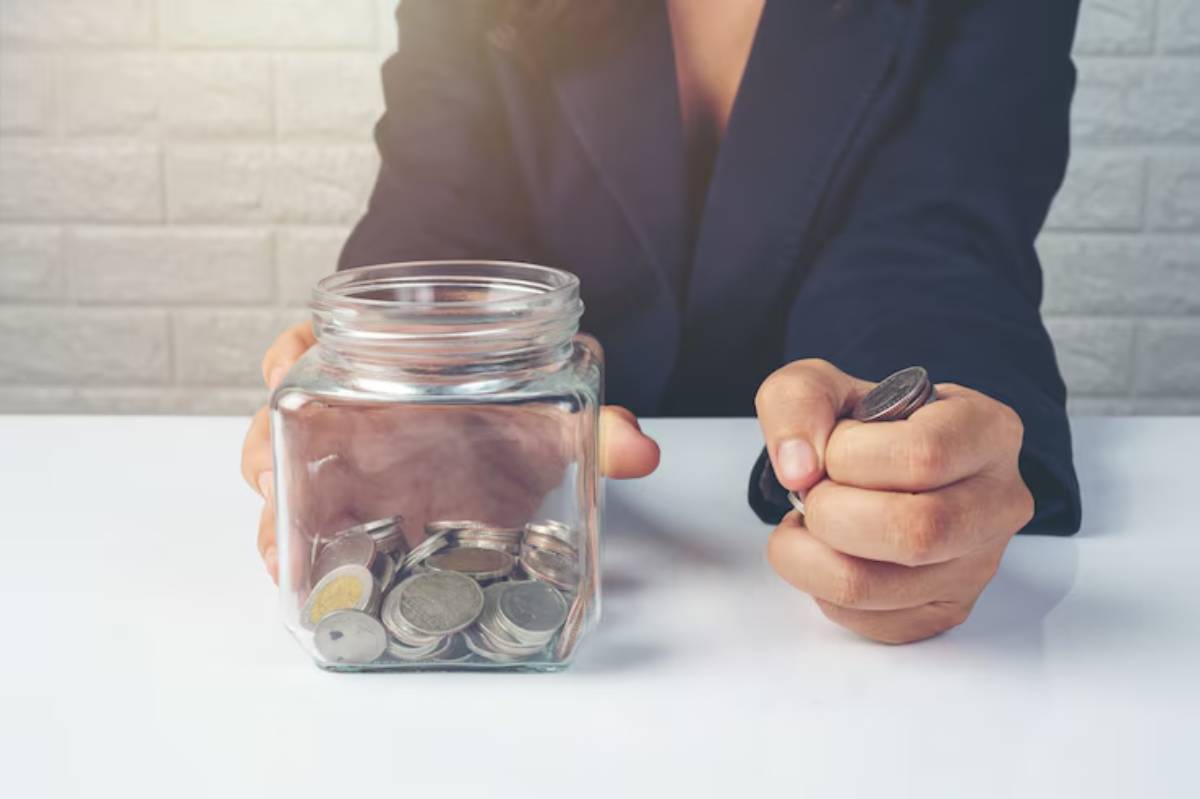 A person in a suit holds a handful of coins, poised over a glass jar filled with more coins, symbolizing saving or budgeting.