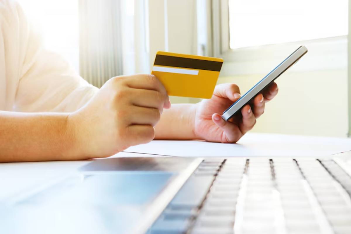A person holds a yellow credit card in one hand and a smartphone in the other, seated at a desk next to a laptop.