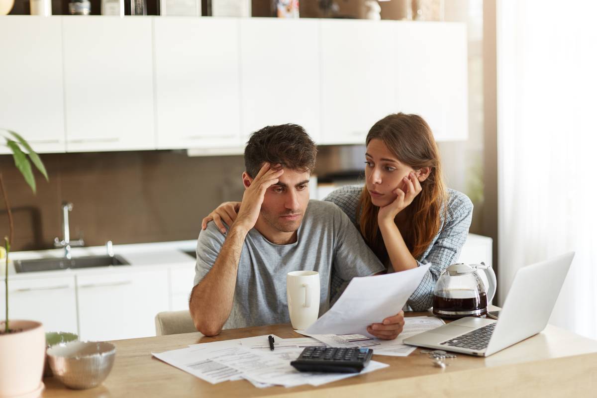 A couple sits at a wooden table covered in documents, a coffee mug, and a laptop, engaged in a discussion about finances. 
