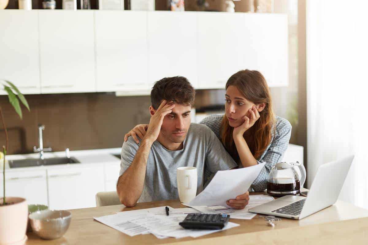 A couple sits at a wooden table covered in documents, a coffee mug, and a laptop, engaged in a discussion about finances.