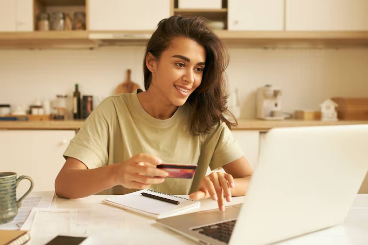 A person sitting at a table with a laptop, holding a credit card, surrounded by notebooks and a coffee mug in a cozy kitchen.