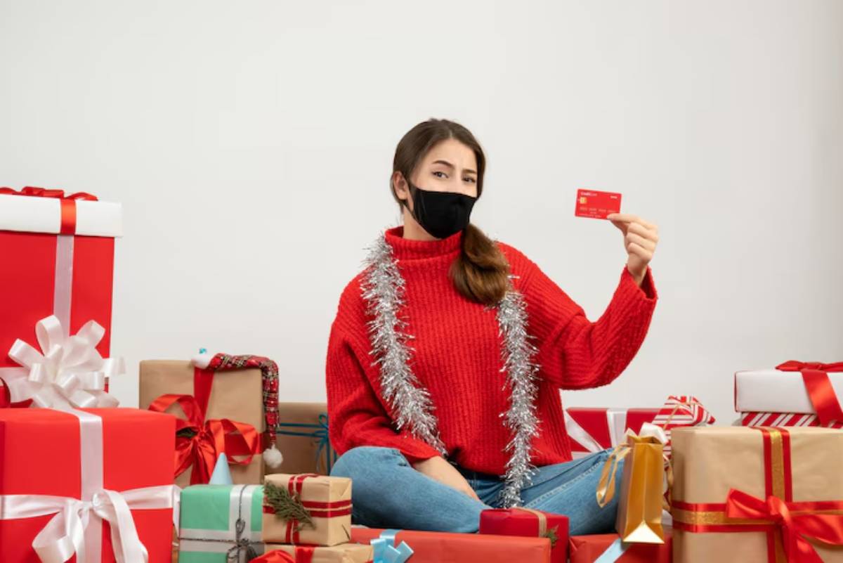 A person in a festive red sweater surrounded by colorful wrapped gifts, holding a gift card with a cheerful expression. 