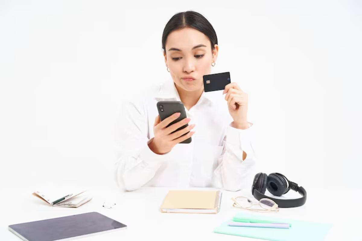 A person in a white shirt holds a smartphone and a card, surrounded by notebooks, glasses, pens, and headphones on a white desk. 